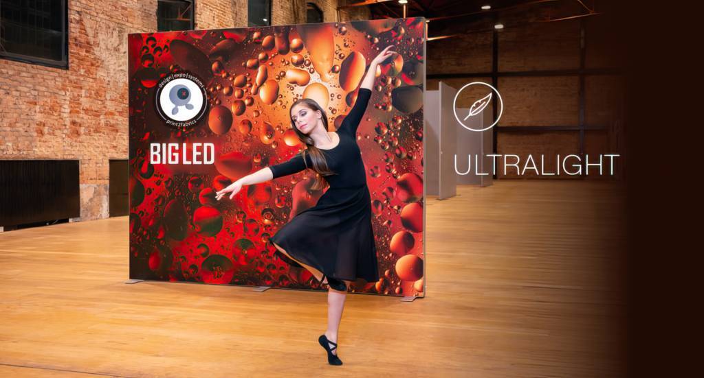 Een strakke tentoonstellingshal met bakstenen accenten vormt het decor voor een opvallende bedrukte BIG LED Lightbox stand met rode bubbeltjesmotieven, aangevuld met een expressieve danseres