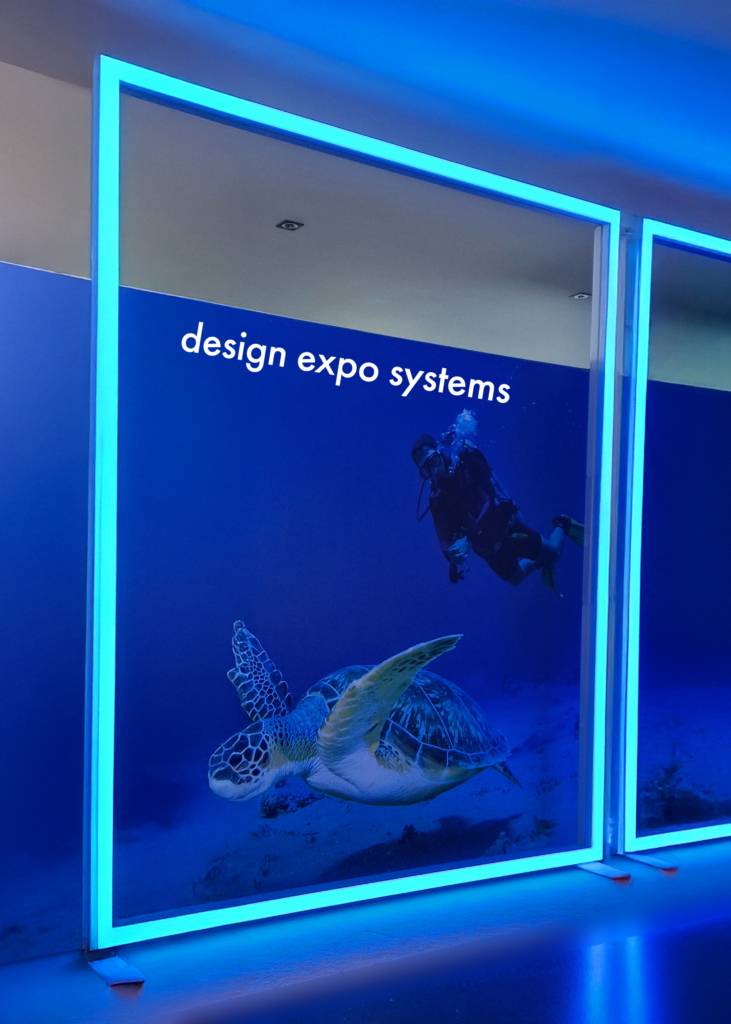 Een groot verlicht display toont een zeeschildpad en een duiker onder water met bovenaan de tekst “design expo systems”. De LineUp RGB SEG Fabric Frame is een hoogwaardig textielframe dat voorzien is van geïntegreerde RGB-verlichting, ideaal voor het creëren van indrukwekkende visuele presentaties.